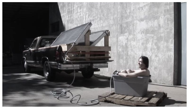 A man bathing in a plastic storage tub on a wooden pallet, using water heated by two of the solar thermal panels from the White House, which are mounted on wooden frames in the back of a pickup truck