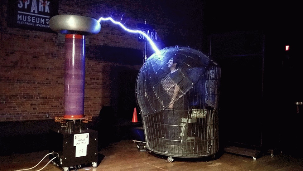 A photo showing a man inside a Faraday cage being protected from a large arcing blue bolt of electricity being produced by a Tesla coil.  